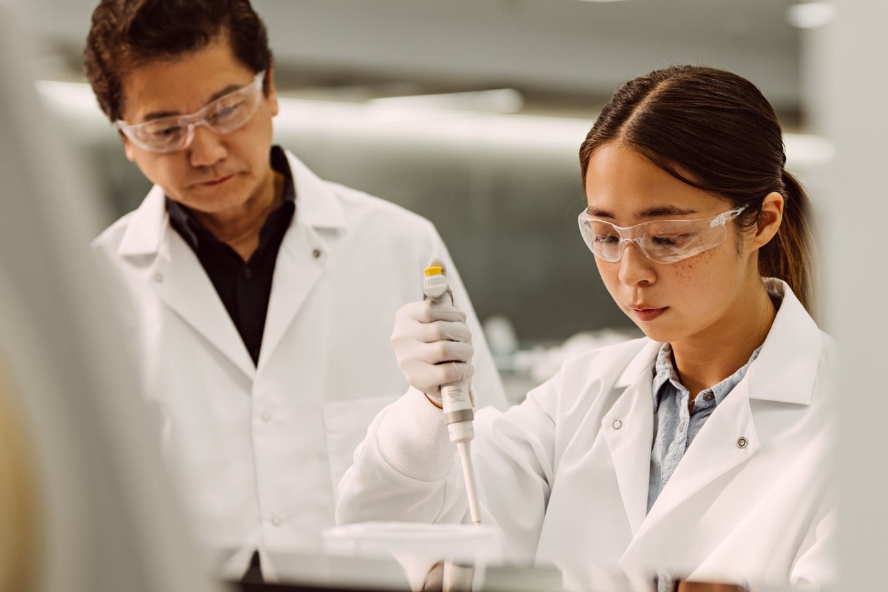 Female is holding a single pipette in one hand, and looking down; male scientist looking over her shoulder observing.