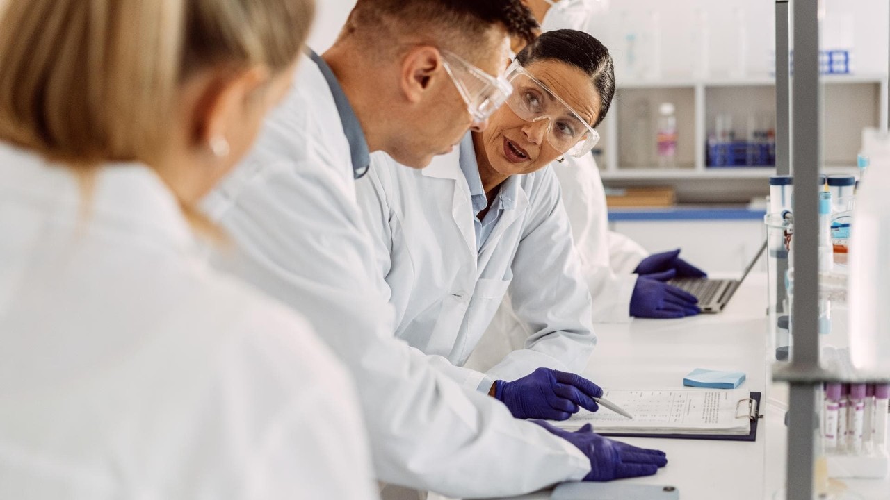 Two scientists, one male and one female, engaging in conversation while the female scientist points to some results on a clipboard; the back of another female scientist, tubes, and other lab equipment blurry in the foreground.